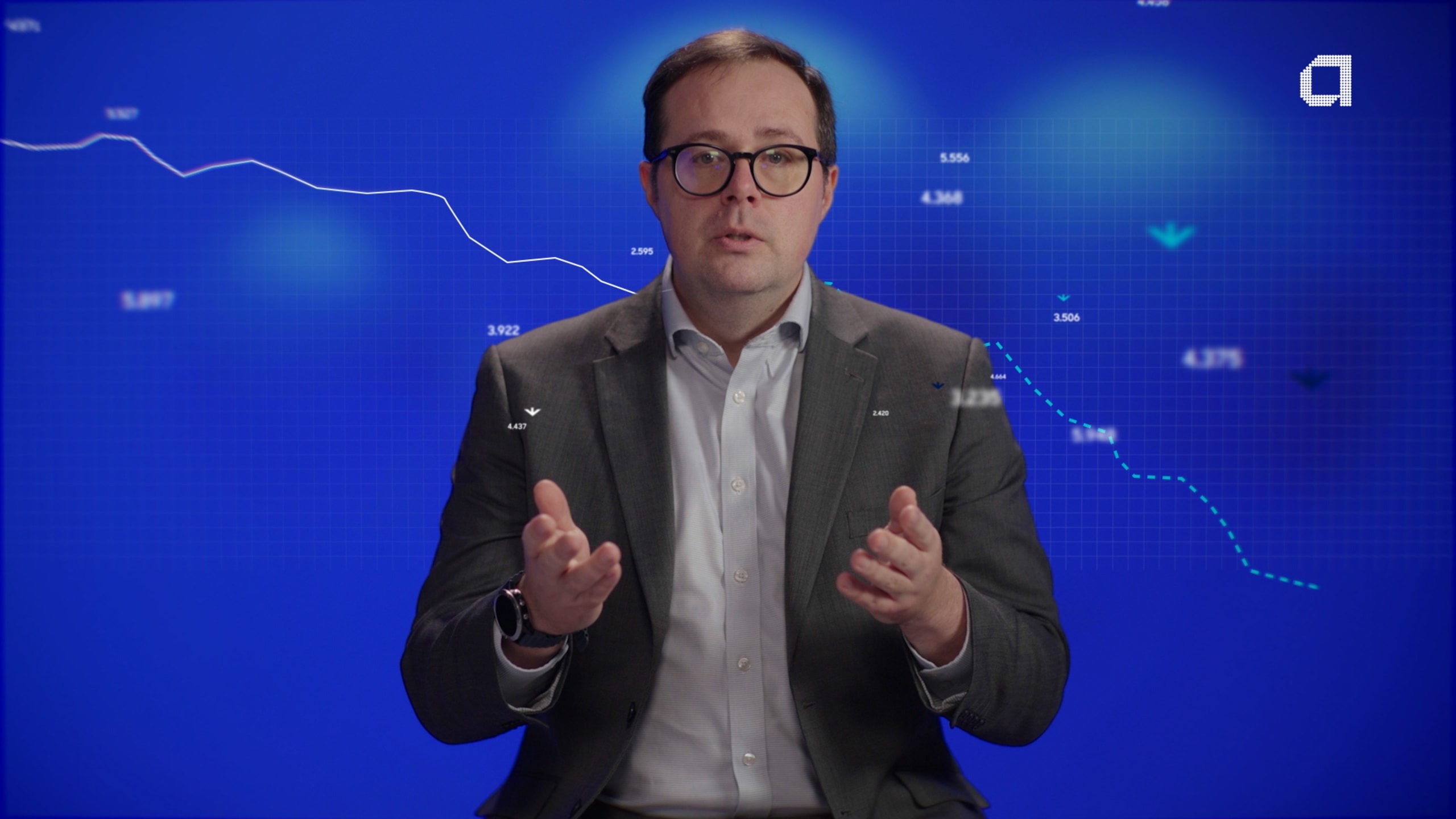 A man in a suit and glasses gestures while sitting. Behind him, a blue background displays a declining line graph with numerical data.