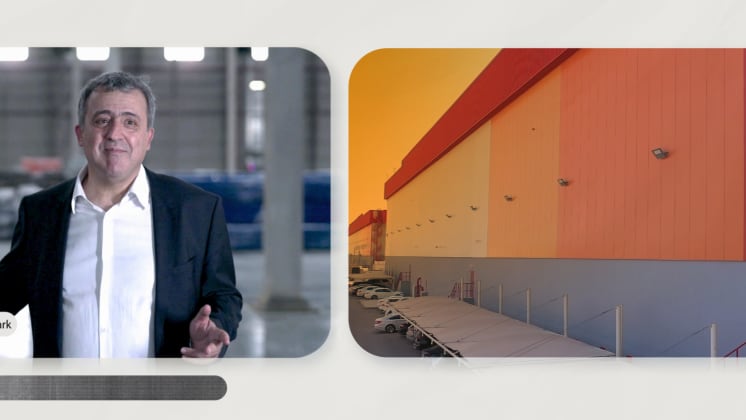 A man in a suit stands indoors, speaking, next to an image of a large warehouse exterior with parked cars.